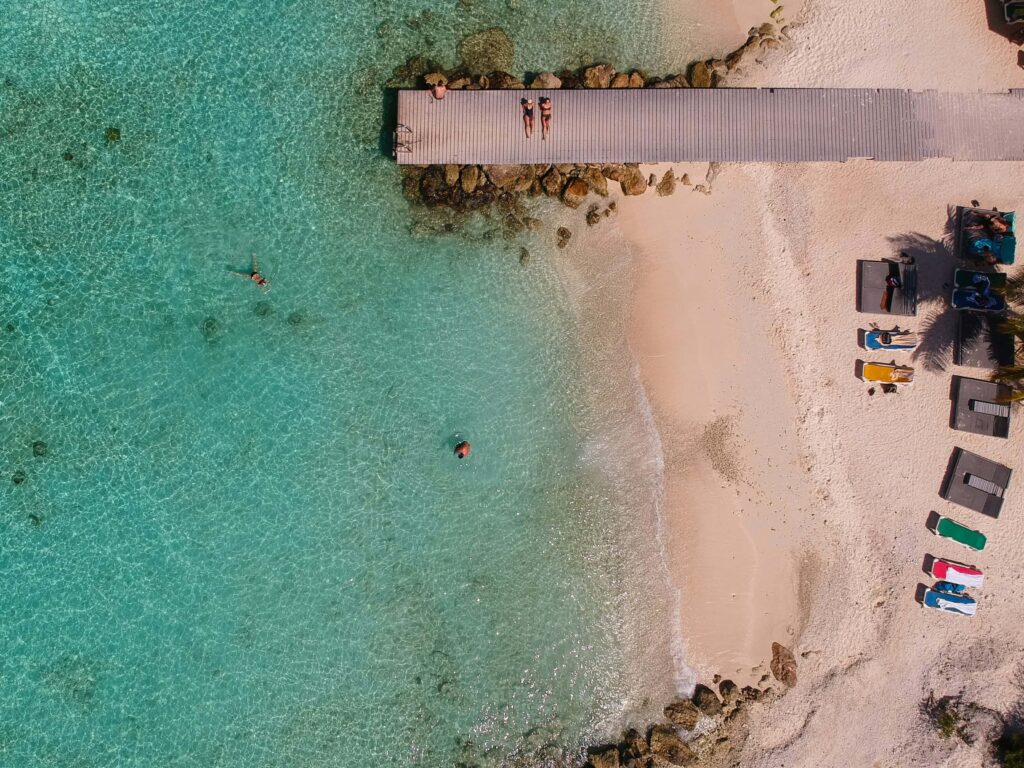 Aerial view of Playa Porto Marie in Curaçao featuring a picturesque bridge over turquoise waters, with tourists relaxing on the sandy beach and enjoying the scenic tropical surroundings.